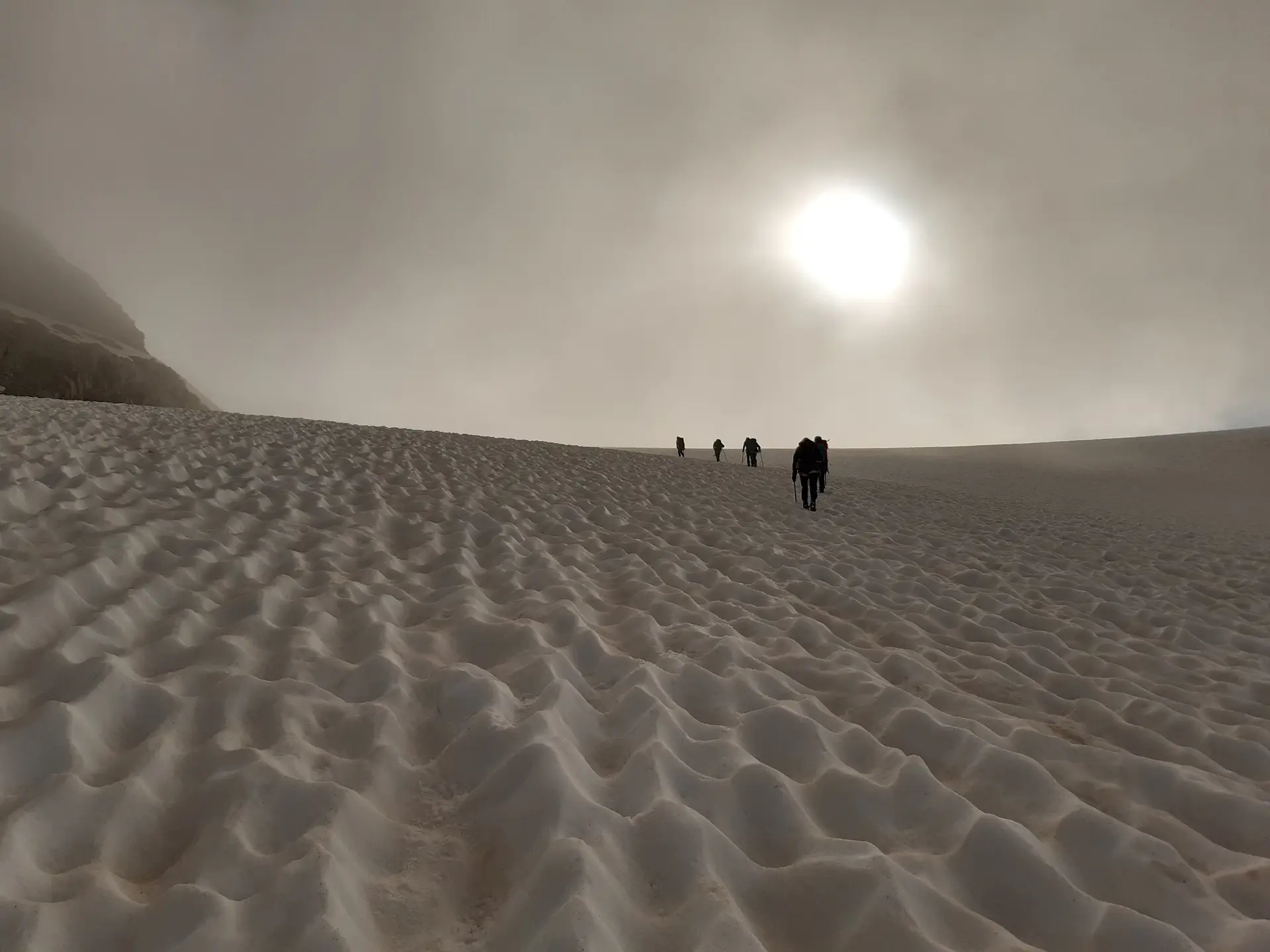 Alpinistes progressant en file sur un névé, vers la lumière du soleil à travers la brume